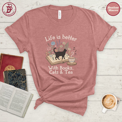A heathered dusty rose short-sleeve t-shirt with a graphic print of a black cat walking over an open book, surrounded by flowers, a teacup, and the text 'Life is better With Books, Cats & Tea'. The shirt is styled in a flat-lay arrangement on a white wood background with books, a coffee cup, and a knot at the bottom hem.