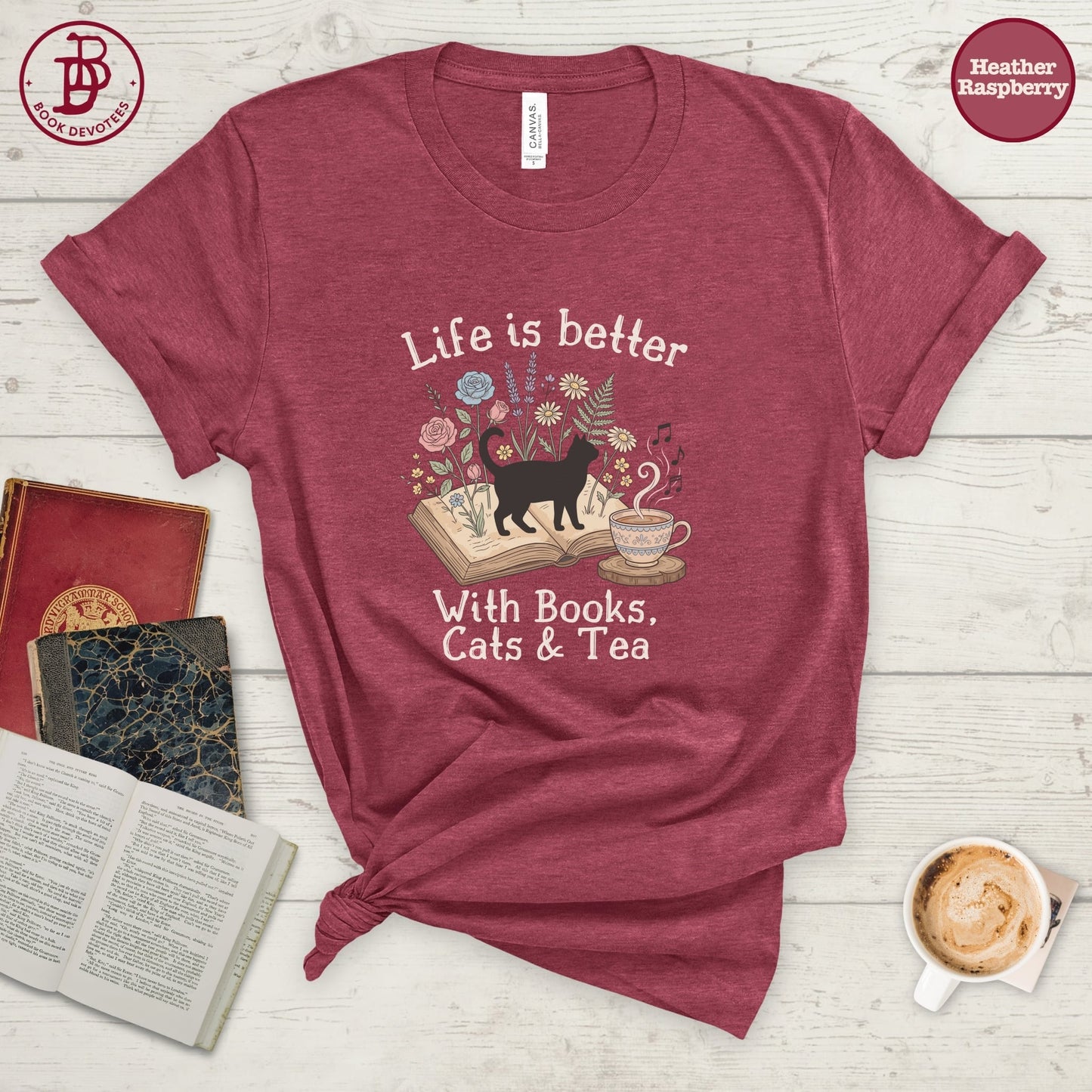 A heathered burgundy t-shirt with rolled sleeves, featuring a graphic of a black cat walking on an open book surrounded by flowers and the text 'Life is better With Books, Cats & Tea'. The shirt is styled in a flat-lay arrangement on a white wood background with books, a coffee cup, and a knotted hem.